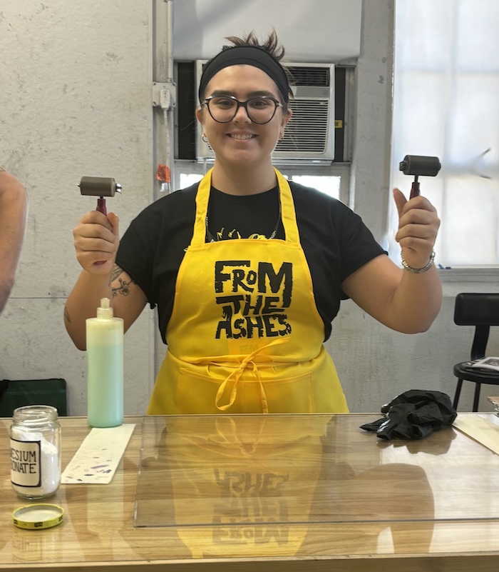 Alex Cox smiles, wearing a bright yellow apron, holding printmaking tools.