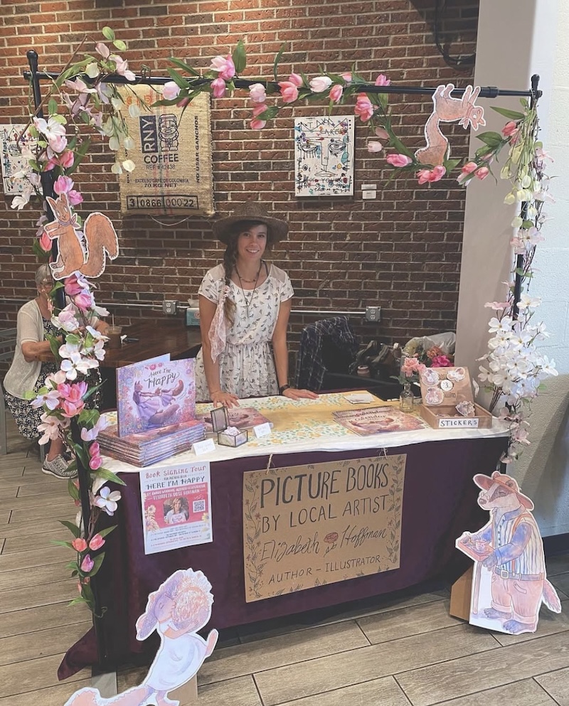 Elizabeth Hoffman smiling behind a stand offering local children's books. The stand is decorated with whimsical flowers and characters.
