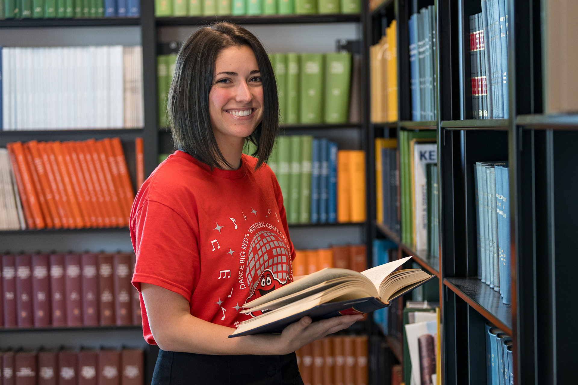 A WKU teacher in a red cardigan leans over a classroom table to help two young female students with their schoolwork; an interactive whiteboard displays a lesson behind them.