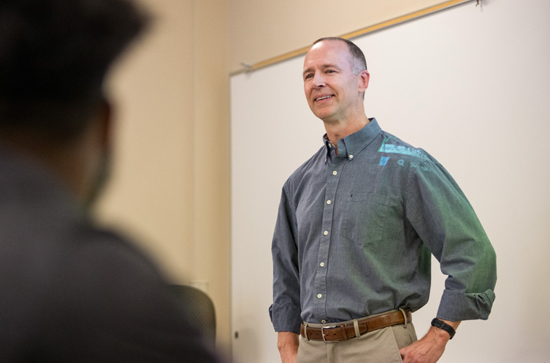 A student researcher monitors a participant's heart rate on a smartphone while the participant holds a plank on a balance board in a psychology lab.