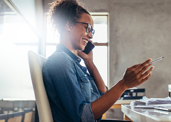 A person sits at a desk, holding a phone to her ear and laughing.