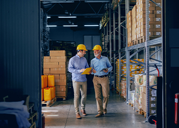 Two people in hard hats walking between large shelves of boxes.