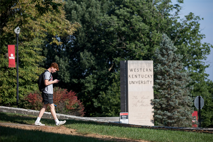 Individual holding a smartphone while walking in front of a concrete statue on WKU's campus