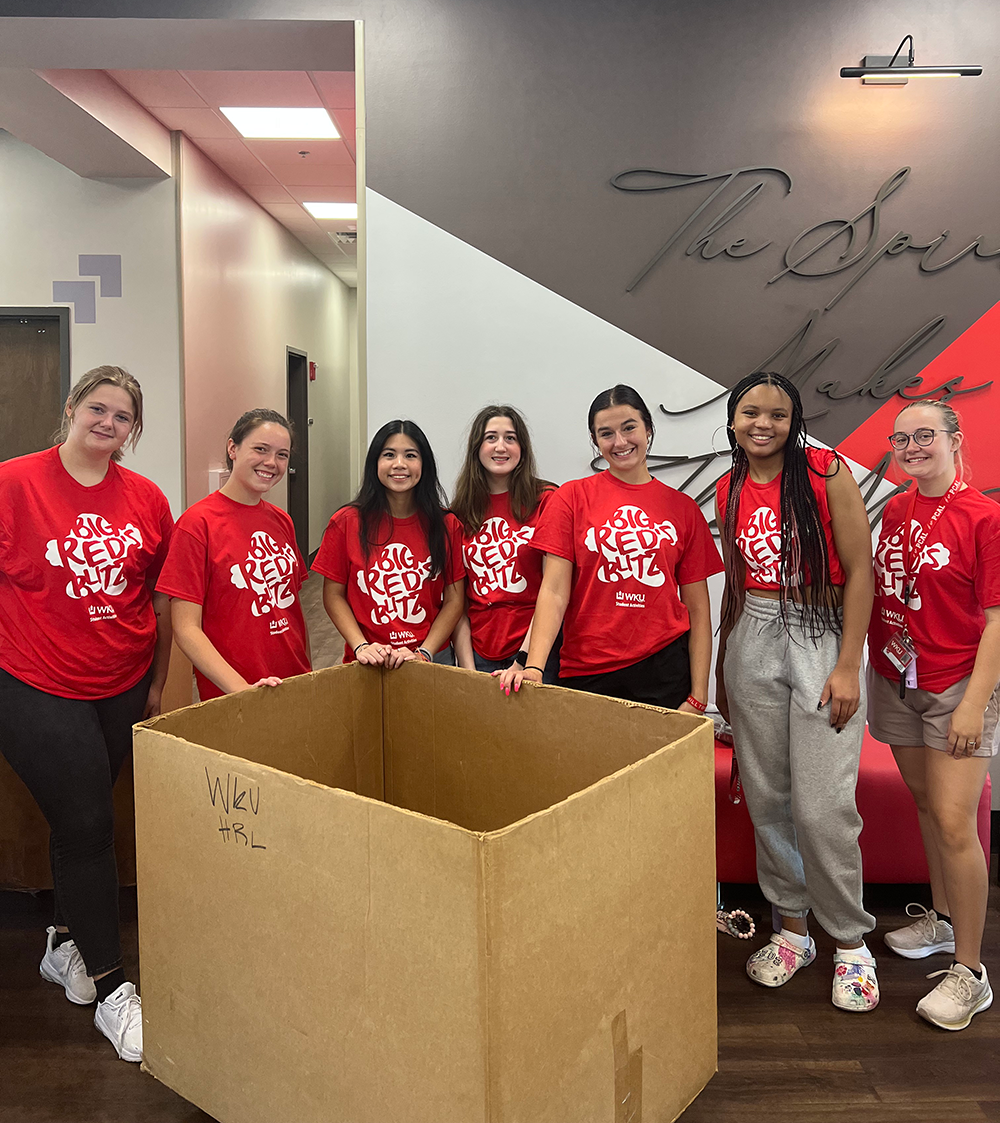 students smiling in front of red/ grey wall with a move in box in front of them