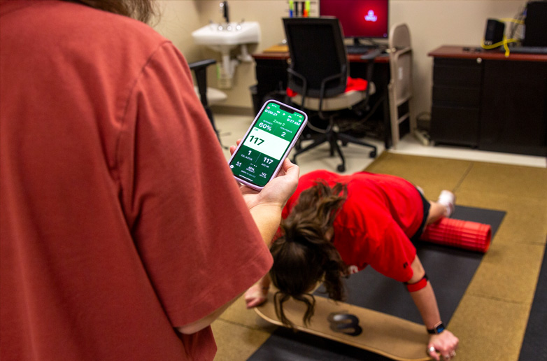 A student researcher monitors a participant's heart rate on a smartphone while the participant holds a plank on a balance board in a psychology lab.
