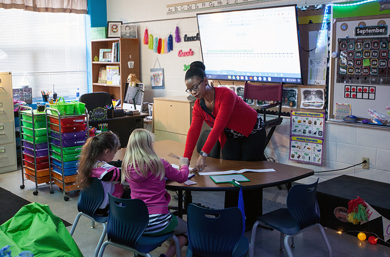 An experienced teacher in a vibrant, modern classroom provides focused, small-group instruction to two elementary students, demonstrating educational leadership in action.
