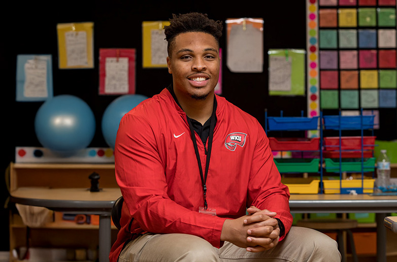 A young male teacher sitting in a bright and colorful elementary school classroom wearing a red WKU zip-up jeacket.